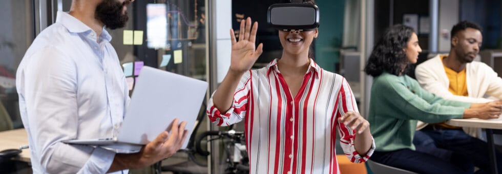 Biracial businesswoman wearing vr headset by caucasian businessman with laptop during meeting. unaltered, business, teamwork, modern office and technology concept.