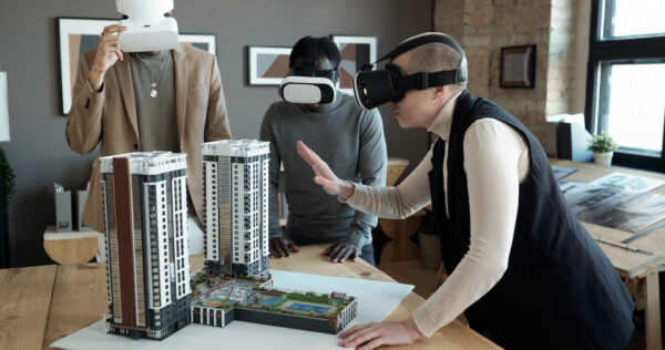 Group of three architects in vr headsets standing by table with layout of new block of flats while one of them making presentation