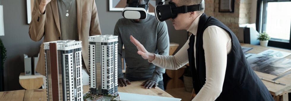 Group of three architects in vr headsets standing by table with layout of new block of flats while one of them making presentation